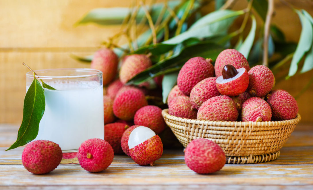 Lichias em uma cesta de palha, ao lado de um copo de suco de lichia, ao fundo, folhas da lichieira