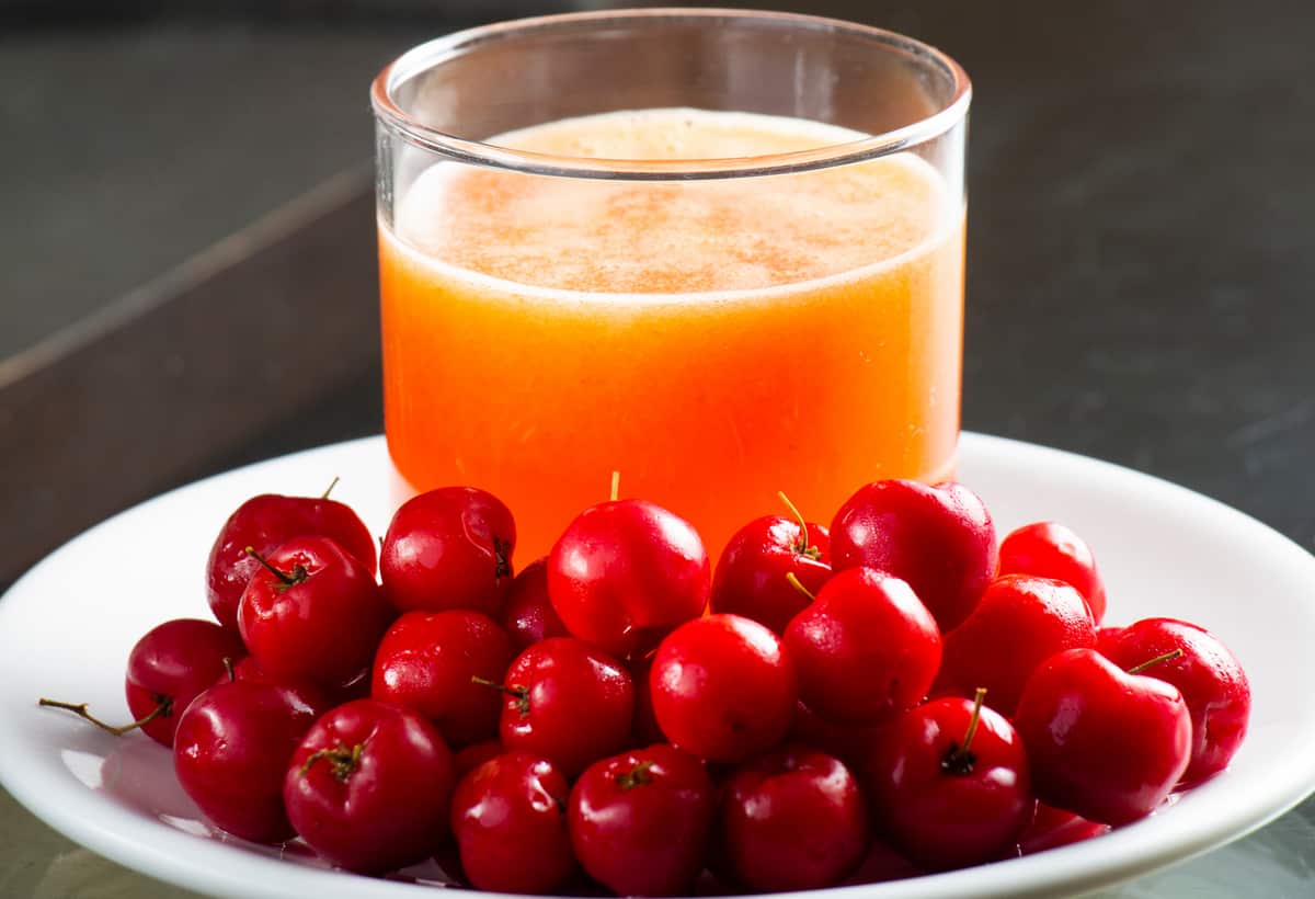 Prato branco cheio de acerolas e com copo de suco de acerola em cima