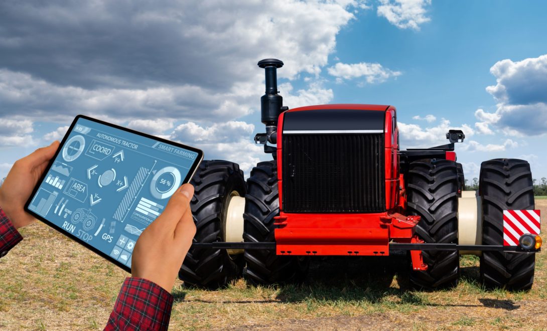 Trator autônomo de cores vermelha e preta em campo. À frente, braços de homem que veste camisa xadrez, segurando tablet com interface de controle.