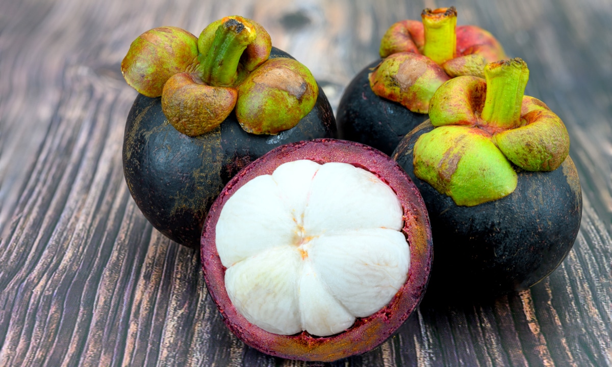 Quatro frutos de mangostaozeiro sobre uma mesa de madeira. Três desses frutos estão inteiros, apresentando casca roxa e cabo em tom de verde e manchas vermelhas. O outro fruto está partido com seis gomos brancos à mostra.