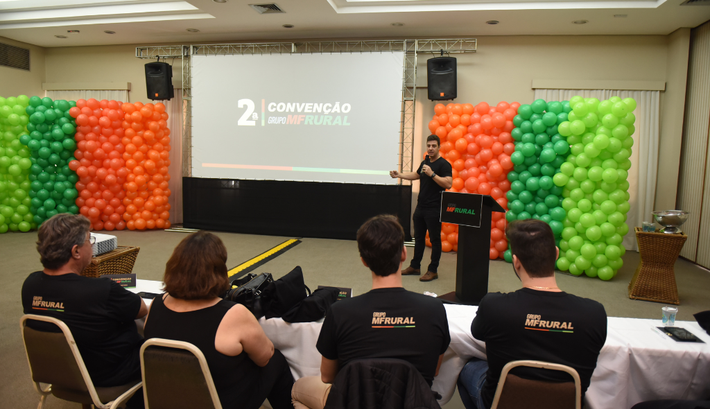 Rafael Lucas palestrando durante a 2ª Convenção do MF Rural. Ao fundo, bexigas das cores verde e laranja e telão com o nome do evento.