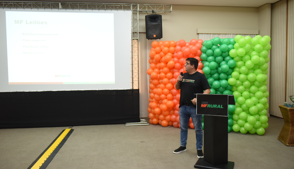 Roberto Lucas palestrando durante Convenção do MF Rural. Ao fundo, bexigas das cores verde e laranja e telão com a apresentação.