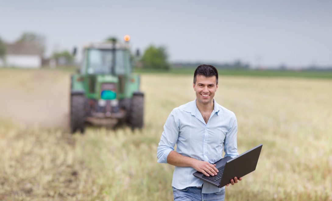 Homem segurando laptop em campo e trator ao fundo