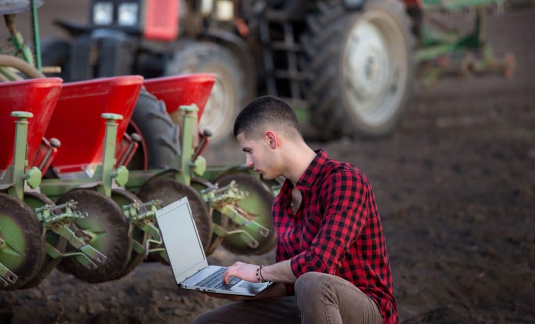 Homem de camisa xadrez vermelha e preta manuseando notebook em frente a trator agrícola.