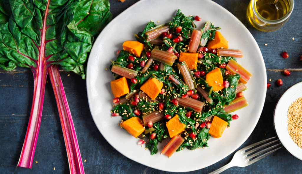 Prato de salada com acelga e outros ingredientes de coloração laranja e vermelha. Do lado esquerdo estão posicionadas folhas de acelga cruas e inteiras e do lado direito um vidro com azeite, sementes de romã e uma espécie de farelo em prato