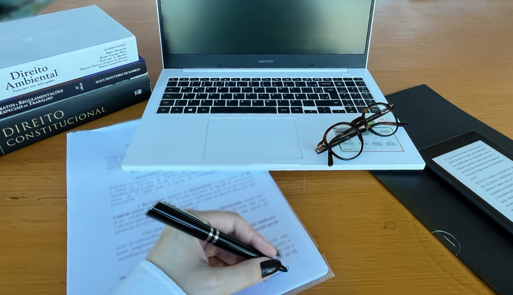 Mão assinando documento em mesa com computador, livros, papéis e Kindle