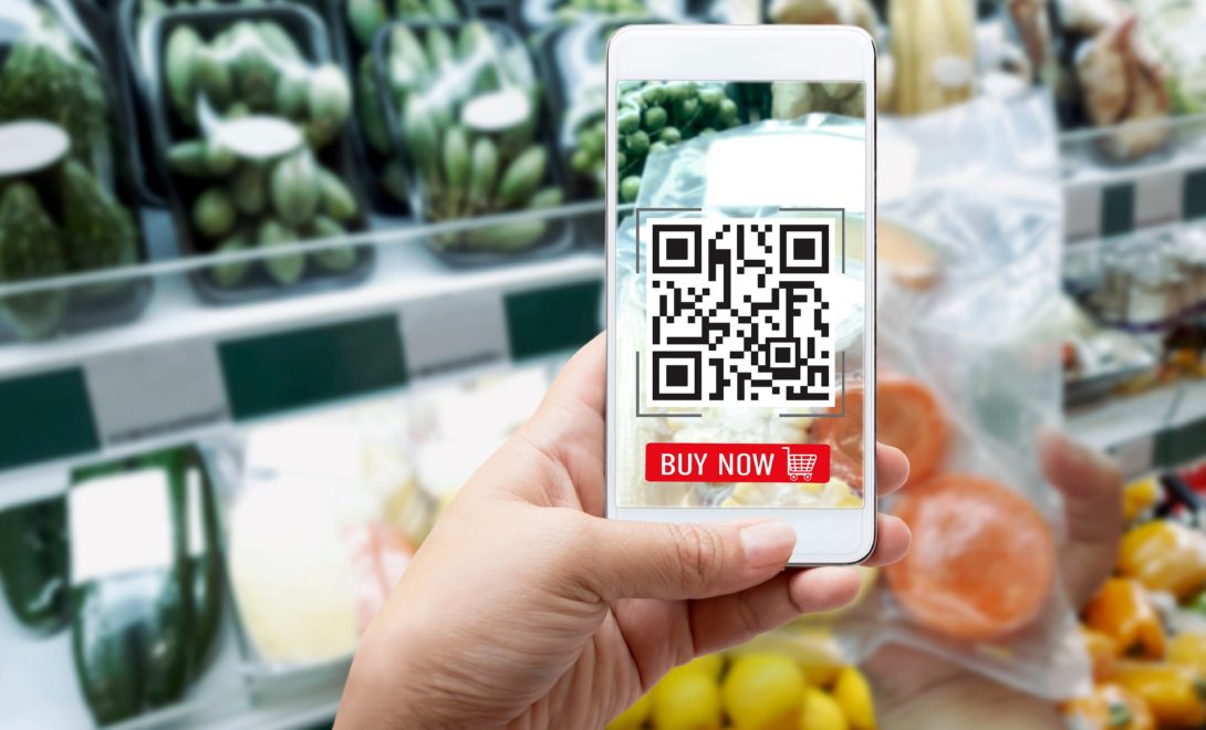 Mão segurando celular apontando para produtos agrícolas embalados em prateleiras de mercado com QR Code na tela