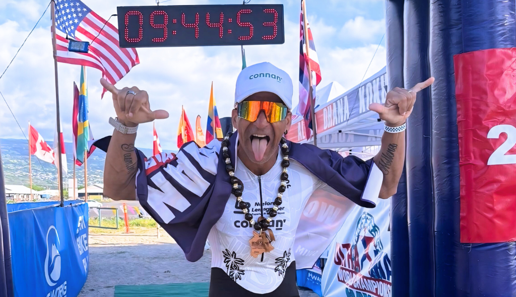 Alessandro Medeiros, atleta carnívoro, em frente à linha de chegada com sua medalha no pescoço e pose descontraída. Ao fundo a marca de sua prova: 9 horas, 44 minutos e 53 segundos