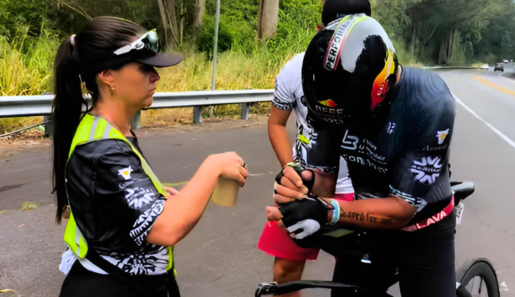 Letícia Moreira, nutricionista, dando eletrólitos ao Alessandro Moreira durante a prova. Alessandro está apoiado na bicicleta enquanto espera que Letícia lhe entregue a bebida