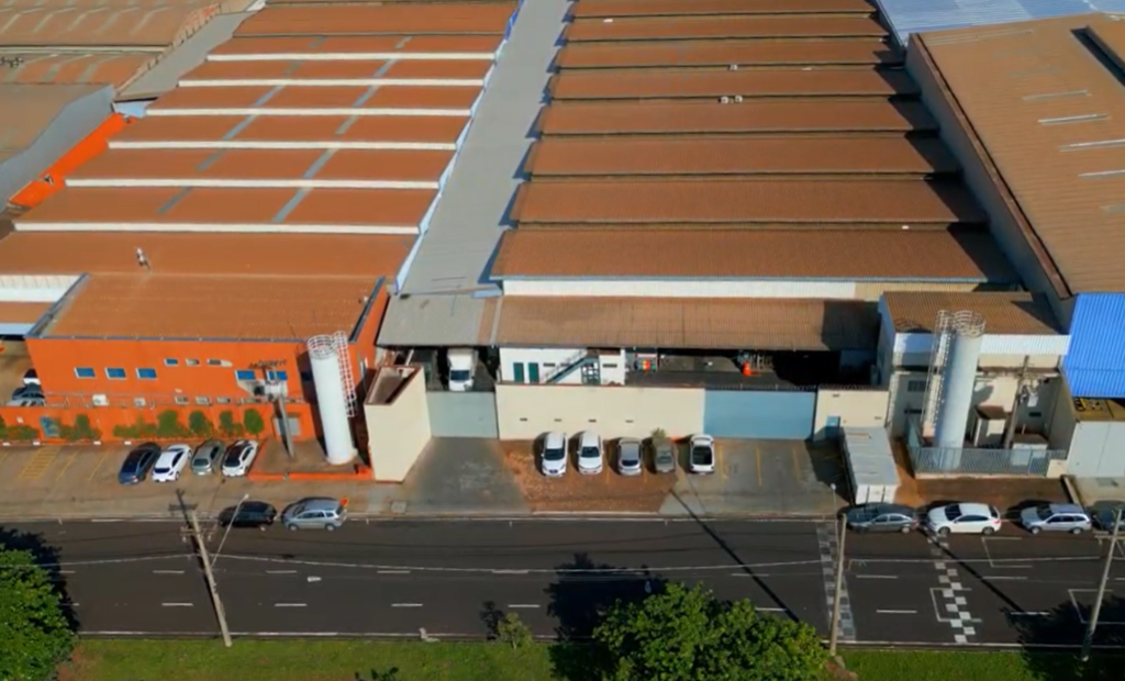 Imagem aérea da fábrica da AGMAQ Agroindustrial, localizada em Ribeirão Preto (SP). A estrutura ocupa uma ampla área com cobertura em telhas metálicas e fachada em tons de laranja, branco e azul. Na frente da fábrica, é possível ver vários carros estacionados e dois grandes silos metálicos, indicando a robustez da operação industrial. A unidade está situada às margens de uma avenida movimentada, com boa infraestrutura urbana ao redor.