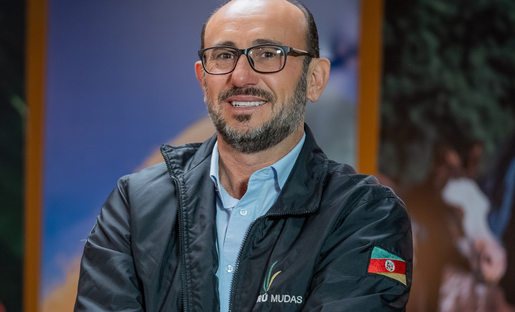 A imagem mostra um homem sorridente, de óculos e barba, vestindo uma jaqueta preta com o logotipo da Xiru Mudas. Na manga da jaqueta, há um patch com a bandeira do estado do Rio Grande do Sul. O fundo está desfocado, mas é possível ver que é um ambiente com painéis ou imagens coloridas, possivelmente um estande ou cenário relacionado ao agro.