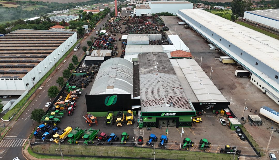 A foto é uma vista aérea das instalações da empresa Sucatão Soluções Agrícolas. O complexo industrial ocupa uma ampla área urbana e exibe uma estrutura robusta, com diversos galpões, pátios e áreas cobertas. Na parte frontal, é possível ver a fachada da empresa com a identidade visual em destaque: o logotipo verde e o nome "Sucatão" em letras brancas sobre fundo verde escuro.

O pátio está repleto de máquinas e equipamentos agrícolas, como tratores, pulverizadores e colheitadeiras, de diversas cores e tamanhos, organizados em fileiras. Ao fundo, há uma grande área onde peças, implementos e máquinas estão armazenadas, indicando o vasto estoque da empresa. Também se observa o fluxo de veículos e caminhões na rua lateral, reforçando o movimento e a operação ativa do local.