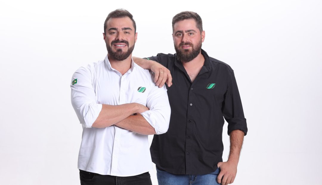 A imagem mostra dois homens em pé, lado a lado, em um fundo branco. Ambos estão sorrindo levemente e demonstram uma postura confiante e amigável. O homem à esquerda veste uma camisa branca com o logotipo verde da empresa bordado no peito e uma bandeira do Brasil na manga. Ele está com os braços cruzados. O homem à direita veste uma camisa preta, também com o mesmo logotipo, e está com uma das mãos no bolso e a outra apoiada no ombro do irmão.