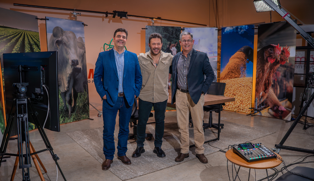 João Bosco no MF Cast Três homens em pé, sorrindo, em um estúdio de gravação com temática do agronegócio. Eles estão posicionados lado a lado, em frente a painéis verticais com imagens de gado, lavoura, grãos e aves. O ambiente conta com mesa, cadeiras, câmeras, iluminação profissional e equipamentos de áudio, indicando a gravação de um programa ou entrevista relacionada ao agro.