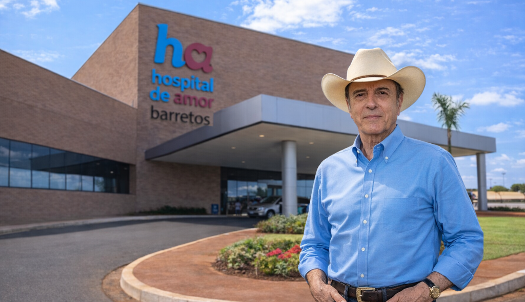 Henrique Prata e Hospital de Amor Homem de chapéu de cowboy e camisa azul está em frente à entrada do Hospital de Amor de Barretos. Ao fundo aparece o prédio moderno do hospital com a fachada de tijolos e o logotipo “ha hospital de amor barretos”. O homem está em primeiro plano, olhando para a câmera, com expressão séria, enquanto o céu azul e a área de acesso ao hospital aparecem ao redor.