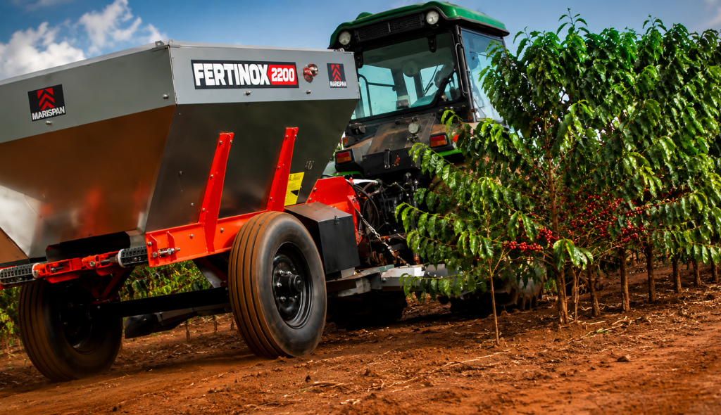 Fertinox Distribuidor de fertilizantes Fertinox acoplado a um trator em operação em lavoura de café, realizando adubação mecanizada entre as linhas do cafezal com foco em tecnologia e eficiência no campo.