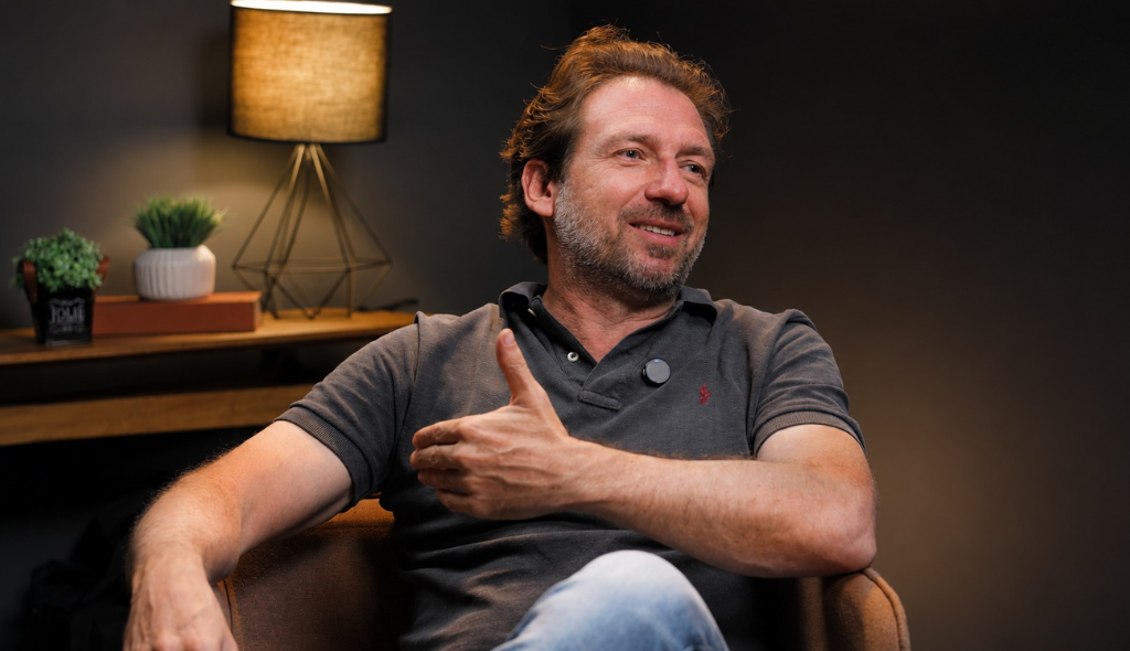 Carlo Barbieri, fundador da SVX Homem sentado em uma poltrona durante a gravação de um podcast, sorrindo e gesticulando enquanto fala. Ele veste uma camisa polo cinza e está em um estúdio com iluminação quente, fundo escuro, uma luminária acesa e plantas decorativas sobre um móvel ao fundo. A imagem transmite um clima de conversa descontraída e entrevista profissional.